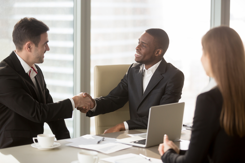 two men shaking hands at a desk in a business office with one woman at the desk