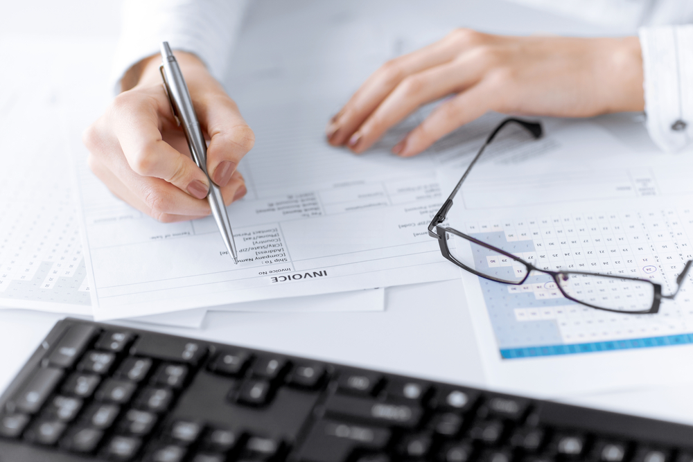person holding pen writing on an invoice document with glasses to the side of the page